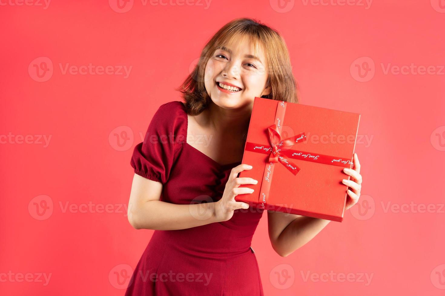 Joven asiática en vestido sosteniendo una caja de regalo roja con expresión alegre en el fondo foto