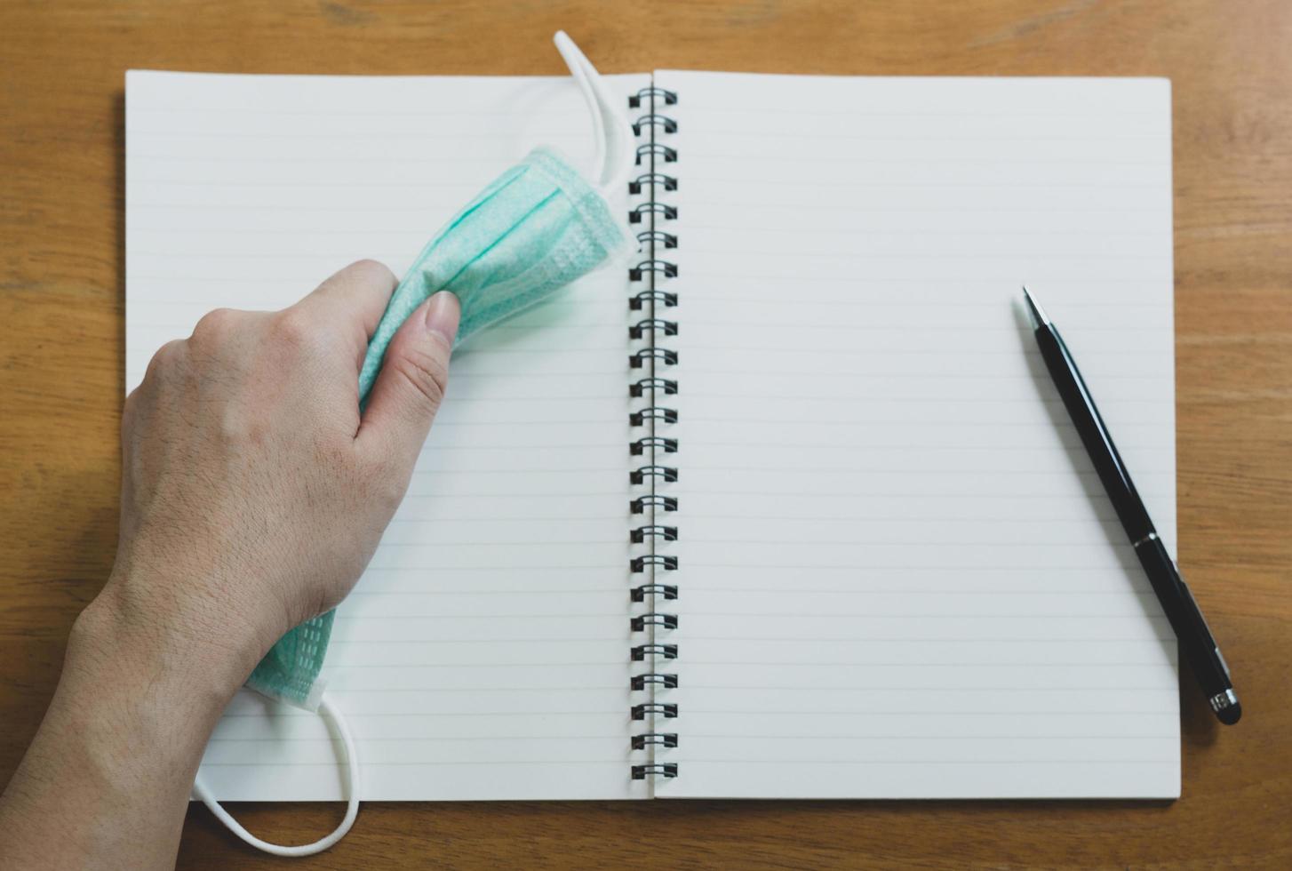 Mano usando la fase de escritura de lápiz en el libro diario con mascarilla quirúrgica en la parte superior de la mesa de madera. covid-19, coronavirus y nuevo concepto de estilo de vida normal foto