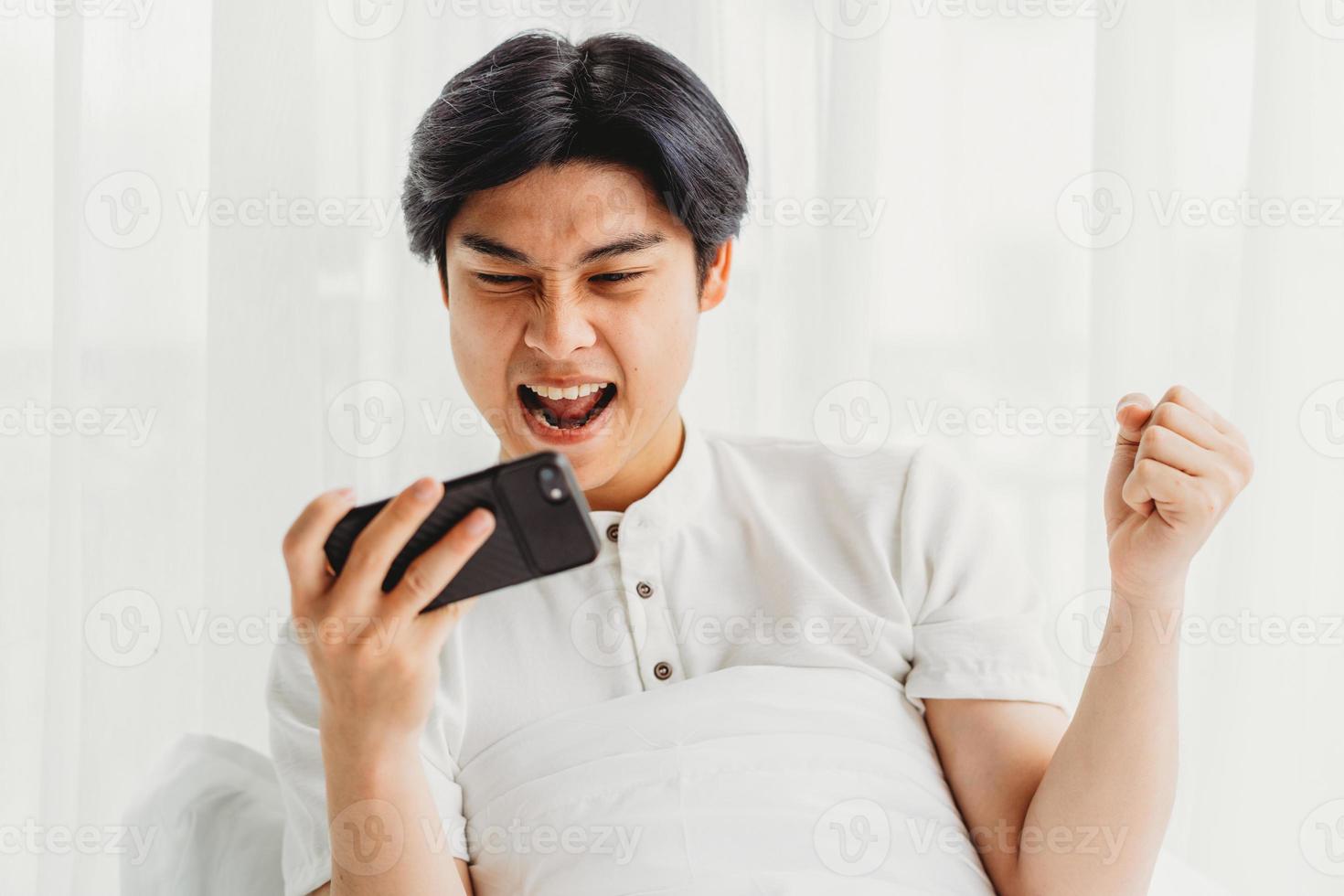 hombre asiático sentado en la cama jugando. el hombre asiático se emociona con la victoria mientras juega el juego móvil foto