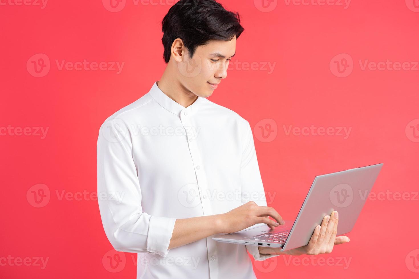 Una foto de un apuesto hombre asiático de pie sobre un fondo rojo con una computadora