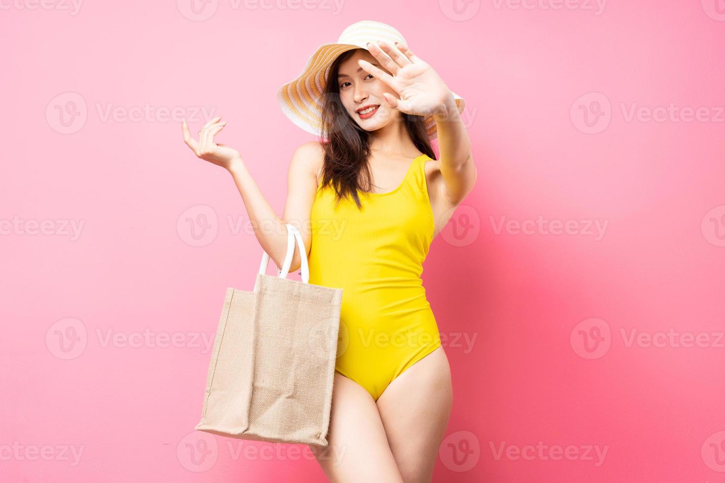 Hermosa joven asiática en traje de baño amarillo, bolso y sombrero de ala ancha posando sobre fondo rosa foto