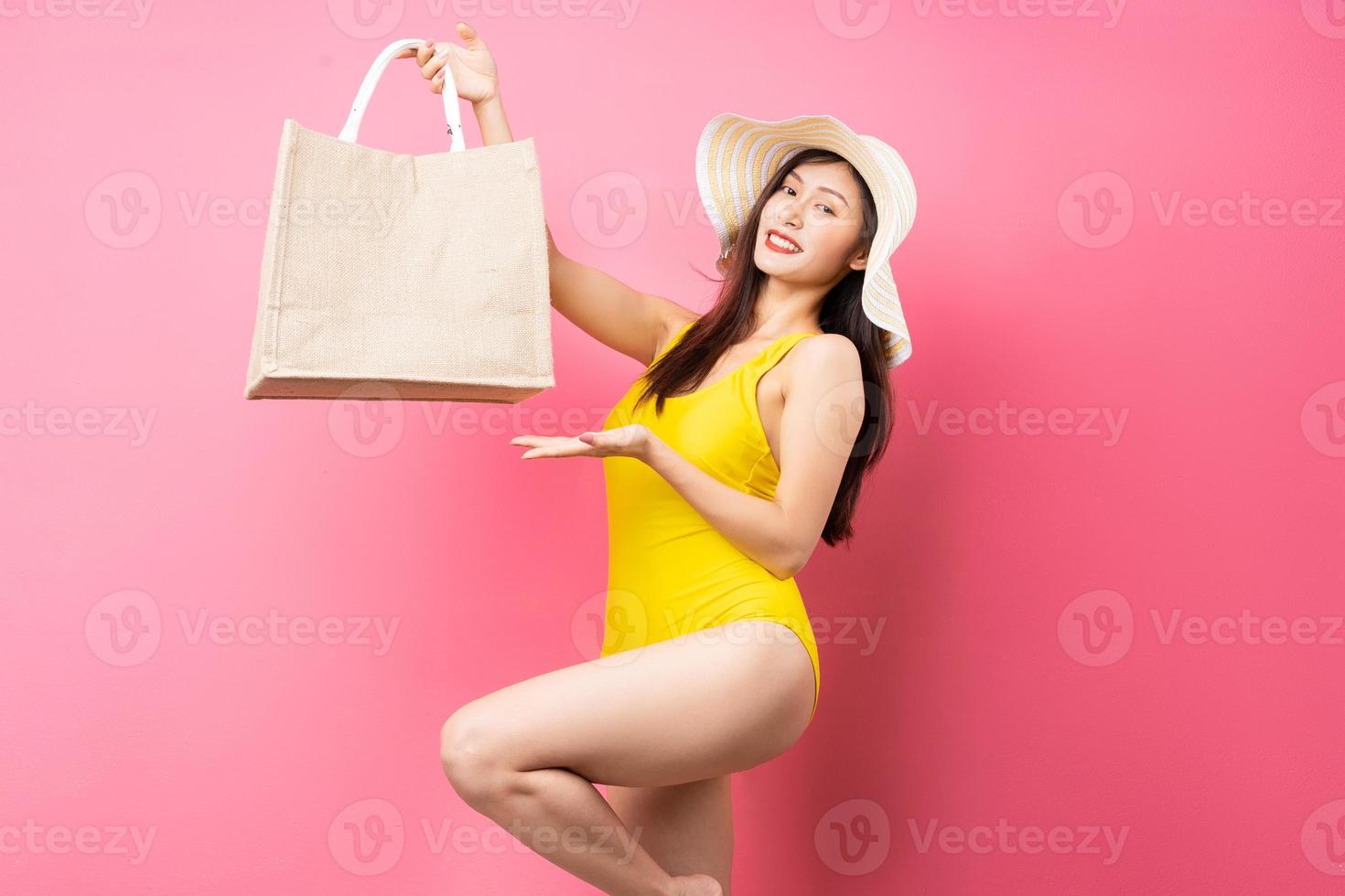 Hermosa joven asiática en traje de baño amarillo, bolso y sombrero de ala ancha posando sobre fondo rosa foto