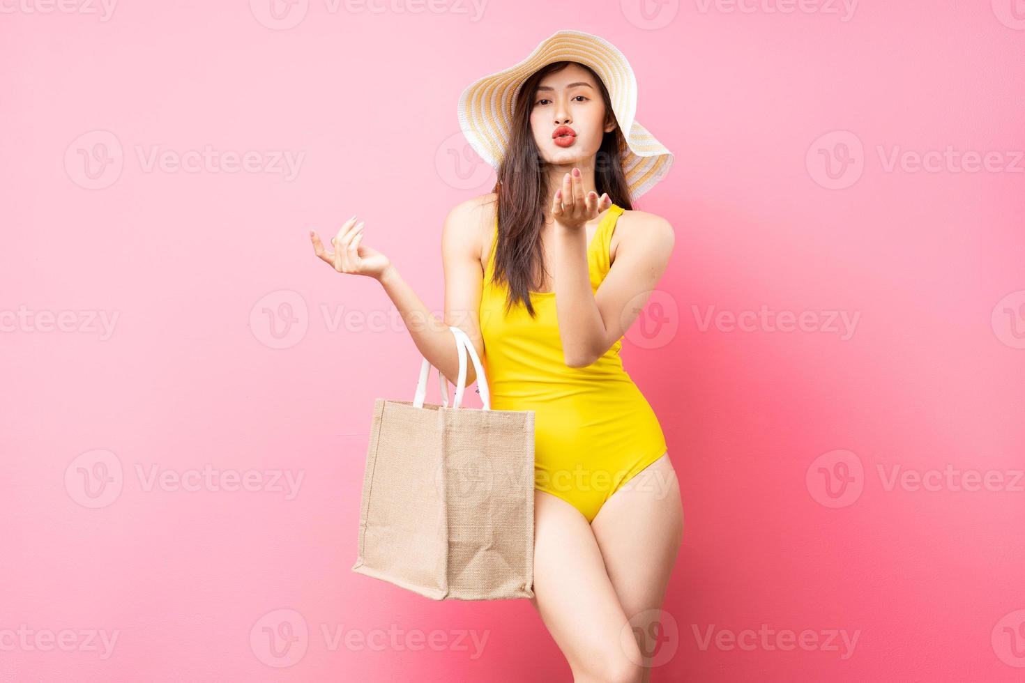 Hermosa joven asiática en traje de baño amarillo, bolso y sombrero de ala ancha posando sobre fondo rosa foto