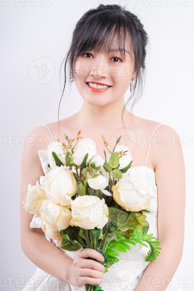 A portrait photo of an Asian bride wearing a wedding dress and smiling beautifully, holding a bouquet of flowers in her hand.