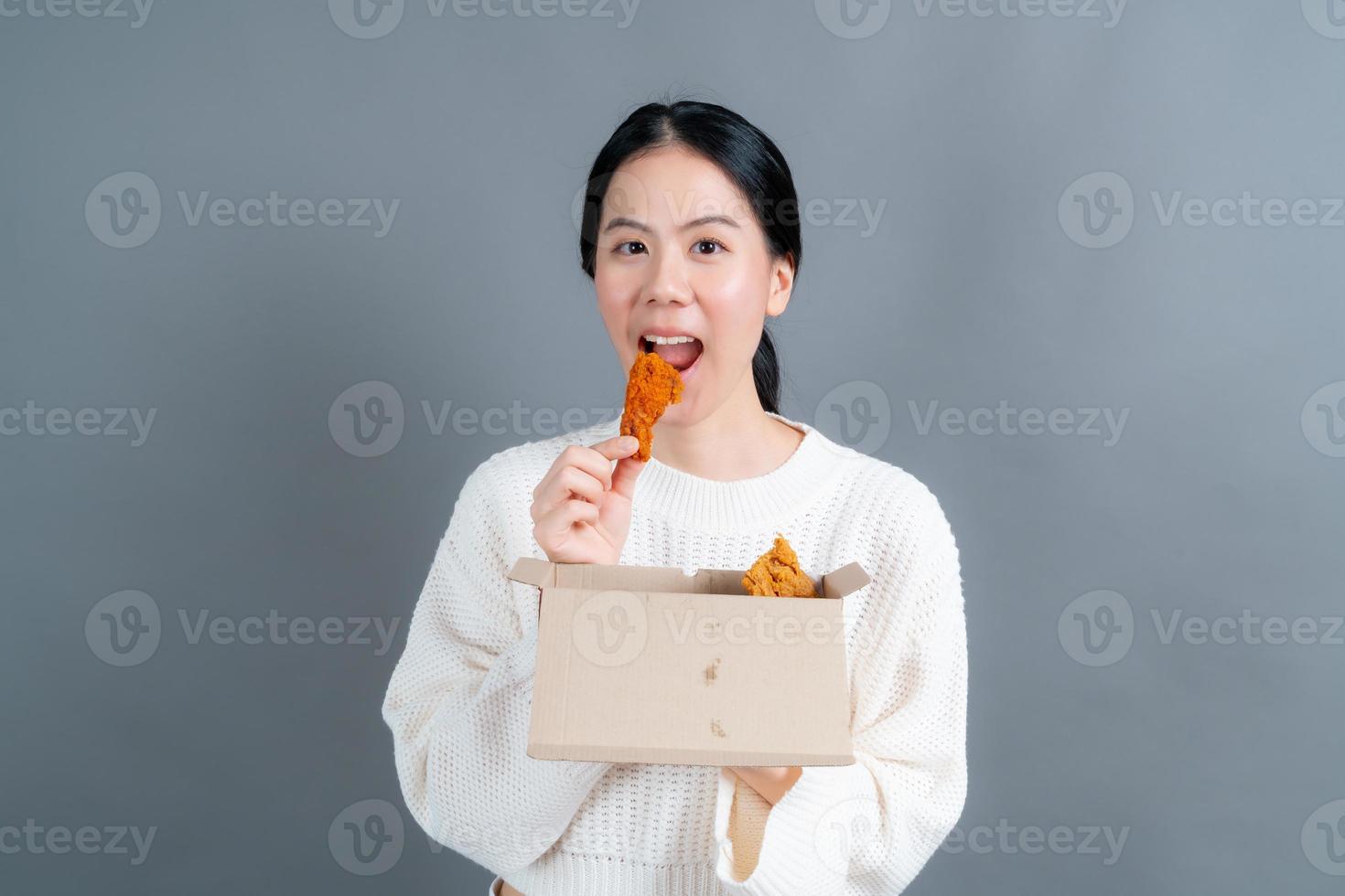 Joven asiática vistiendo un suéter con una cara feliz y disfruta comiendo pollo frito sobre fondo gris foto