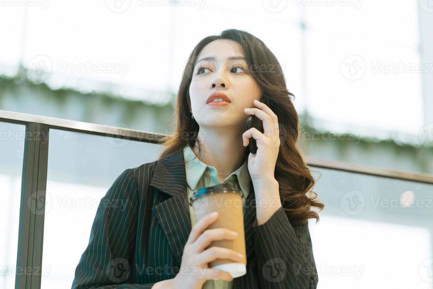 Retrato de una joven directora asiática de pie, tomando café y escuchando el teléfono foto