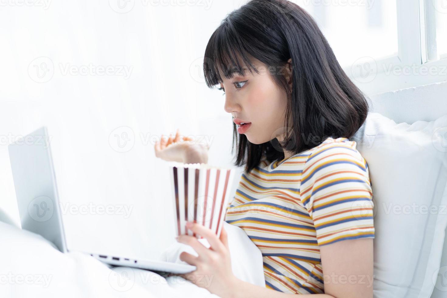 Joven mujer asiática con ordenador portátil para ver programas de televisión, comer palomitas de maíz en casa foto