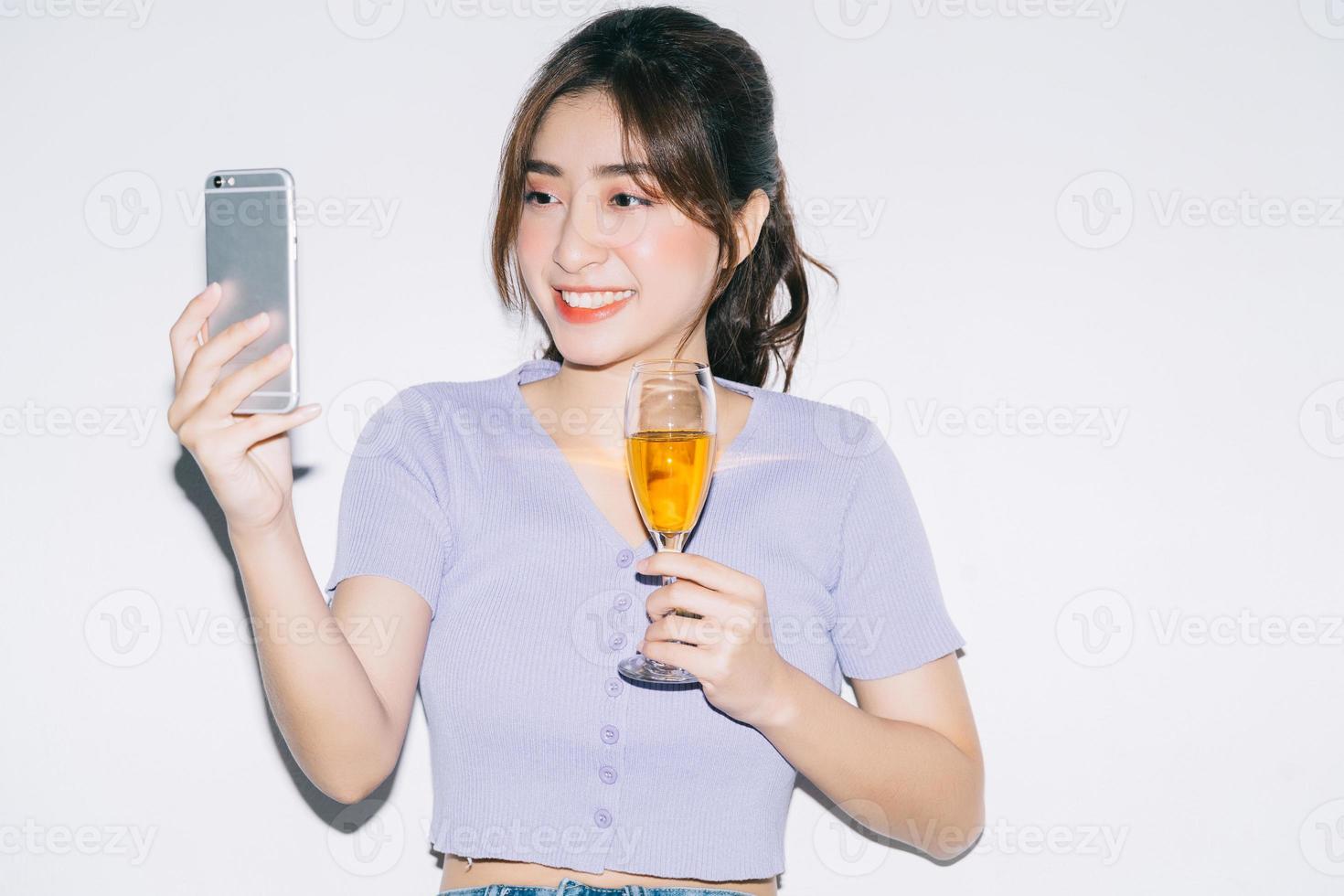 Joven mujer asiática bebiendo vino y con smartphone sobre fondo blanco. foto