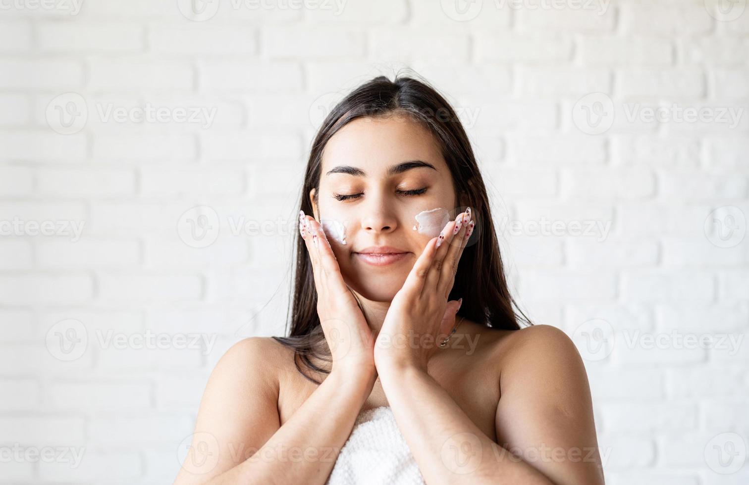 Feliz hermosa mujer vistiendo batas de baño aplicando crema facial en su rostro foto
