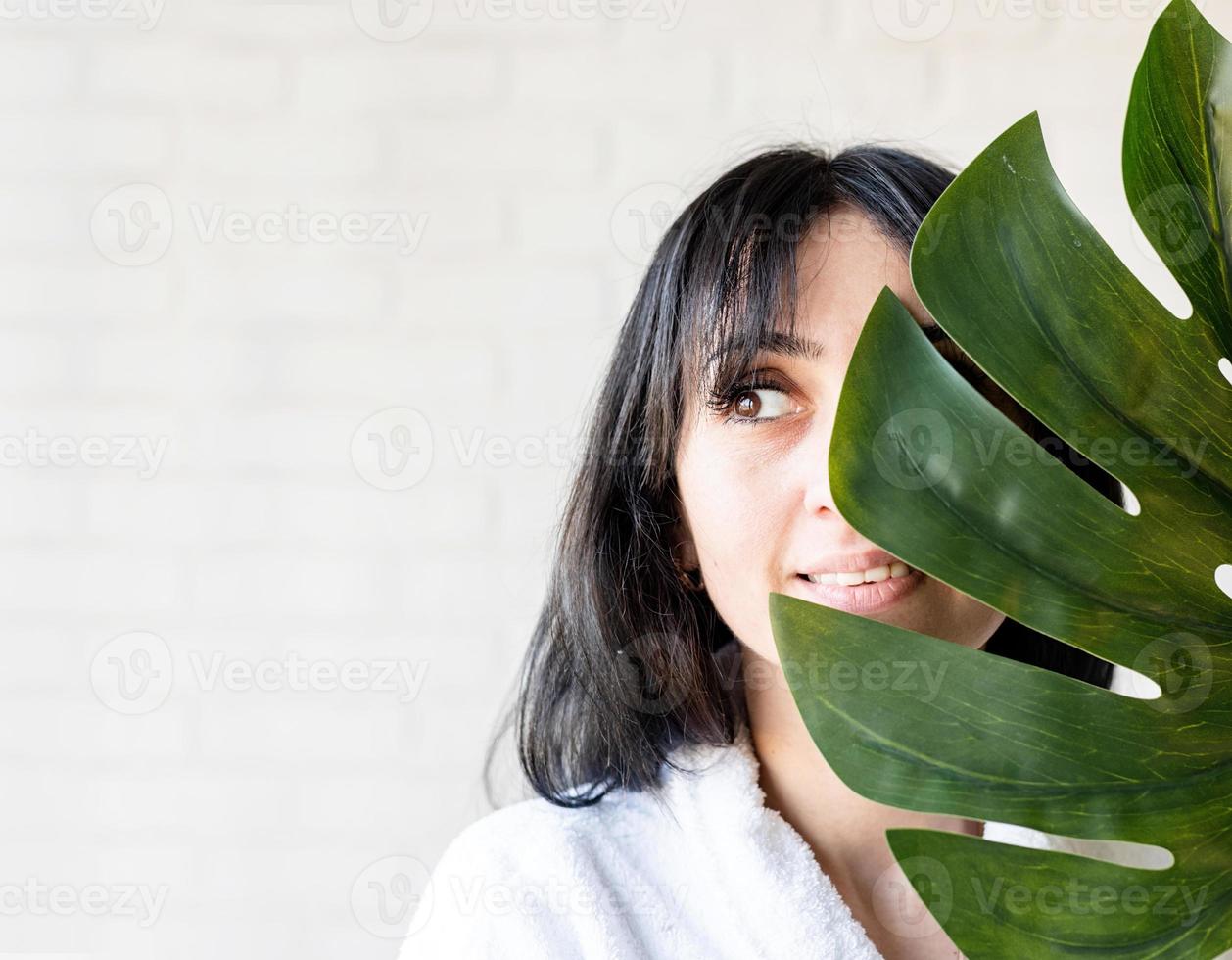 Feliz hermosa mujer de Oriente Medio vistiendo toallas de baño sosteniendo una hoja de monstera verde delante de su rostro foto