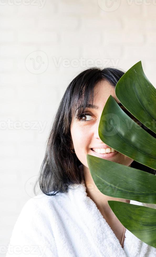 Happy beautiful middle eastern woman wearing bath towels holding a green monstera leaf in front of her face photo