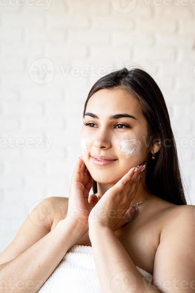 Happy beautiful woman wearing bath robes applying facial cream on her face photo