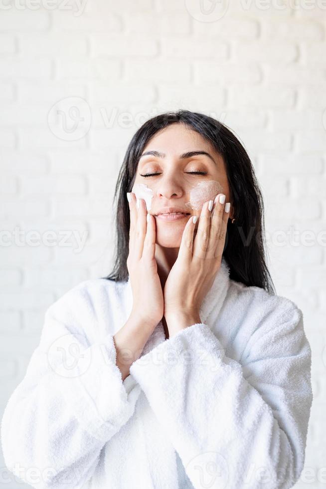 Feliz hermosa mujer de oriente medio vistiendo toallas de baño aplicando crema facial en su rostro foto