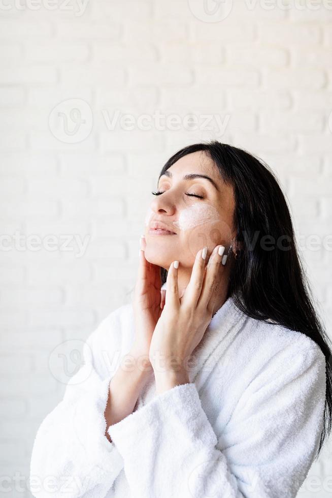 Feliz hermosa mujer de oriente medio vistiendo toallas de baño aplicando crema facial en su rostro foto