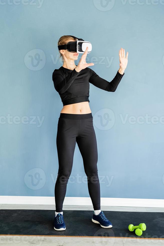 Young blonde woman in sport clothes wearing virtual reality glasses standing at fitness mat using VR interactive menu photo