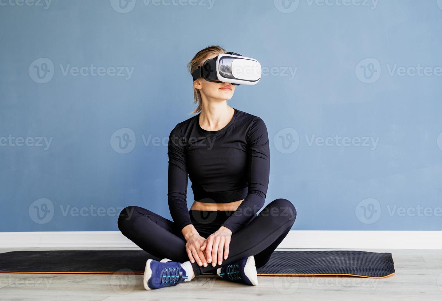 Young blonde woman in sport clothes wearing virtual reality glasses sitting at fitness mat at at home photo