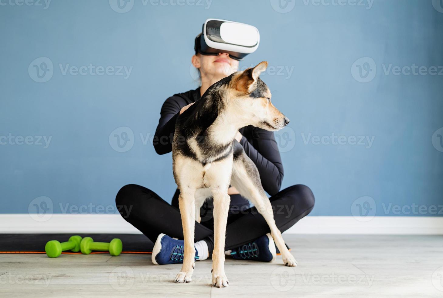 Young woman in sport clothes wearing virtual reality glasses sitting on fitness mat with dog photo