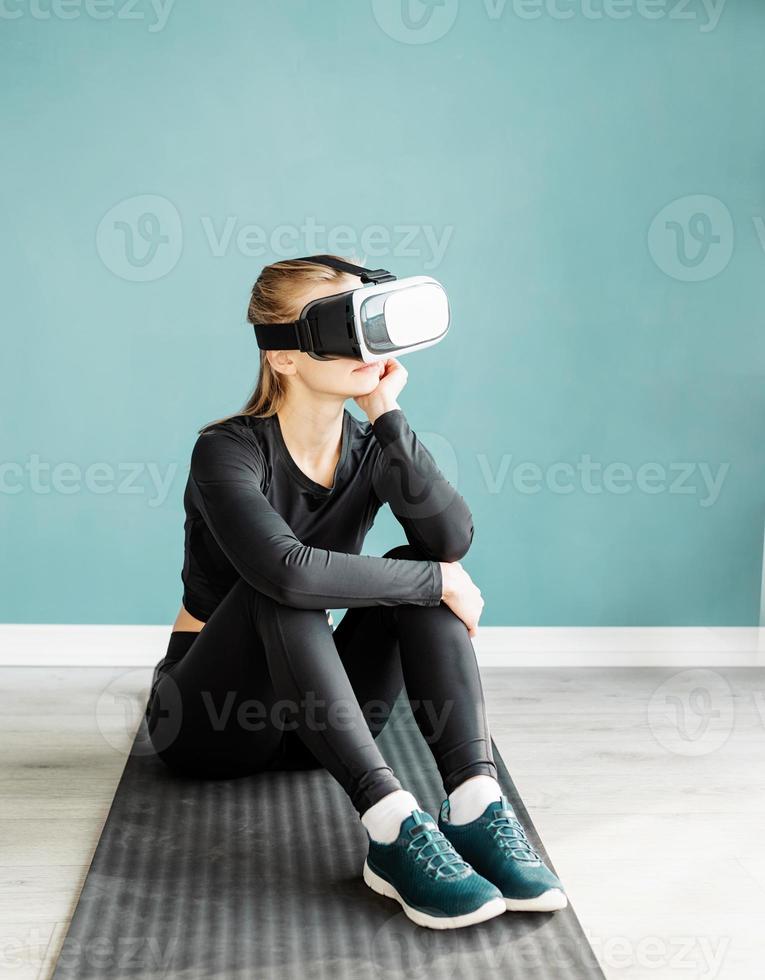 Young blonde woman in sport clothes wearing virtual reality glasses sitting on fitness mat photo