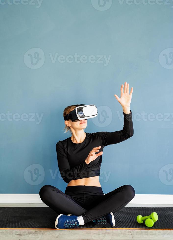Joven mujer rubia en ropa deportiva con gafas de realidad virtual sentado en la estera de fitness utilizando el menú interactivo vr foto