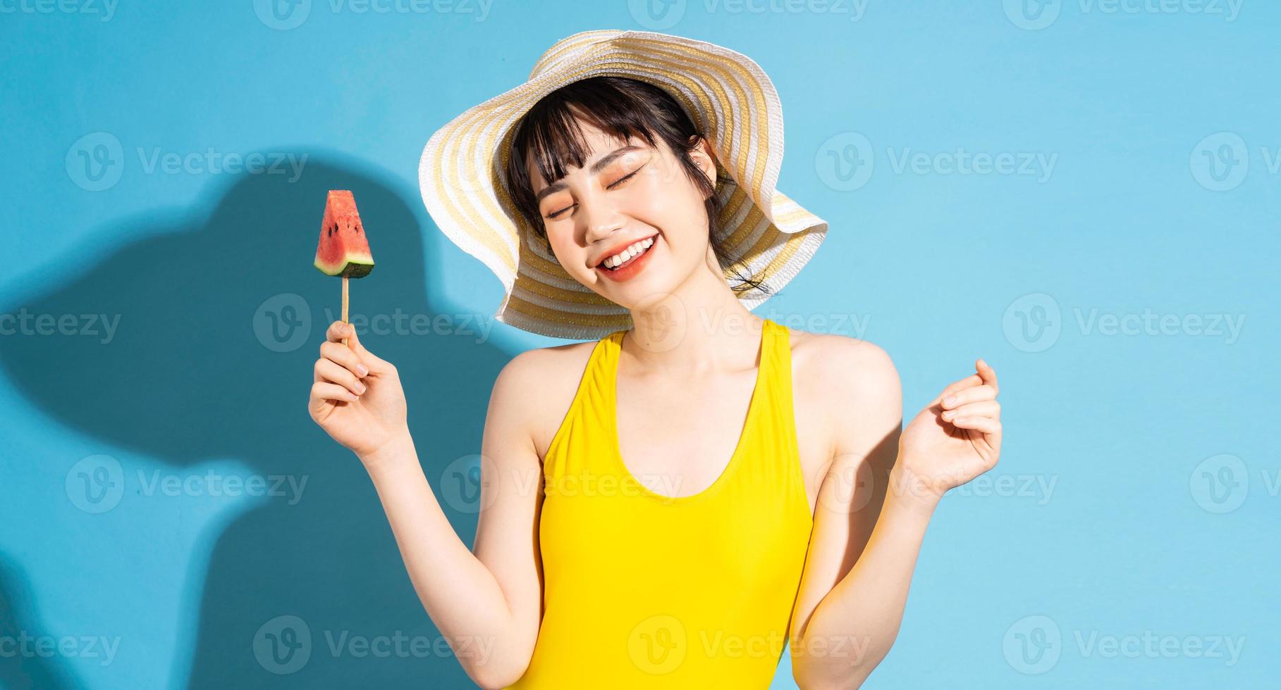 Beautiful Asian woman wearing yellow jumpsuit on blue background and eating tropical fruits, summer concept photo