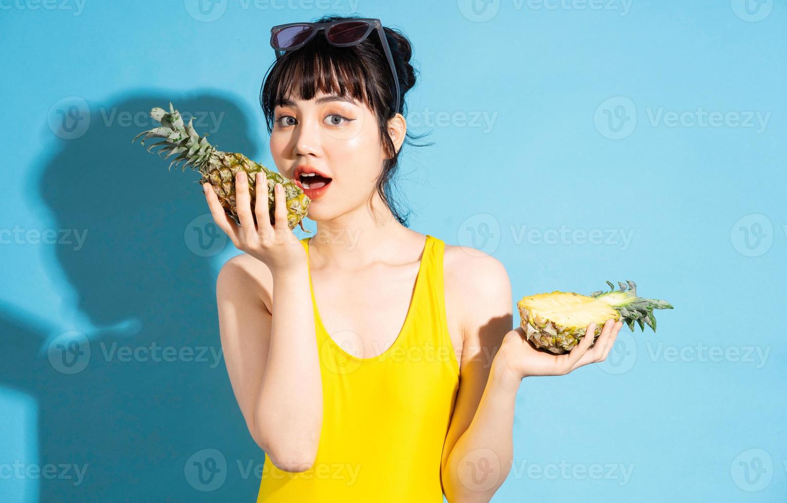 Hermosa mujer asiática vistiendo un mono amarillo sobre fondo azul y comiendo frutas tropicales, concepto de verano foto