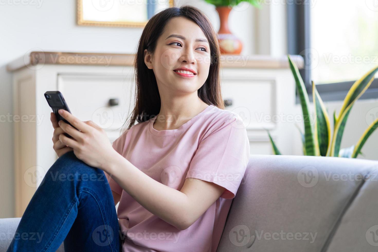 Beautiful young Asian woman sitting on sofa using her phone and looking out the window photo