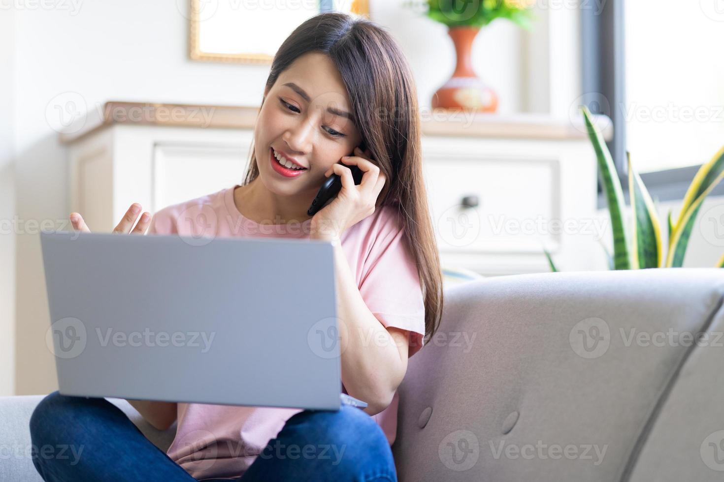 Hermosa joven asiática está llamando y usando la computadora mientras está sentada en el sofá en casa foto