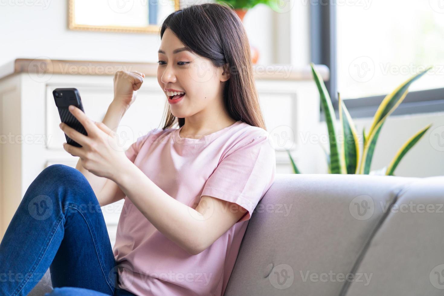 Beautiful young Asian woman sitting on sofa and using phone with victory expression photo