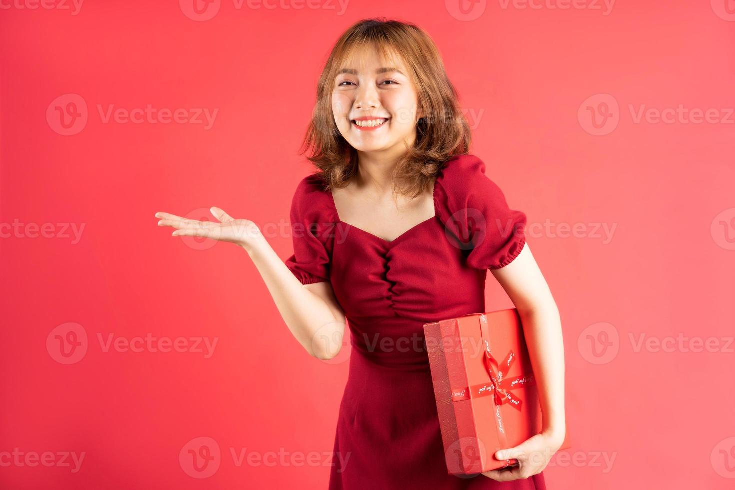 Joven asiática en vestido sosteniendo una caja de regalo roja con expresión alegre en el fondo foto