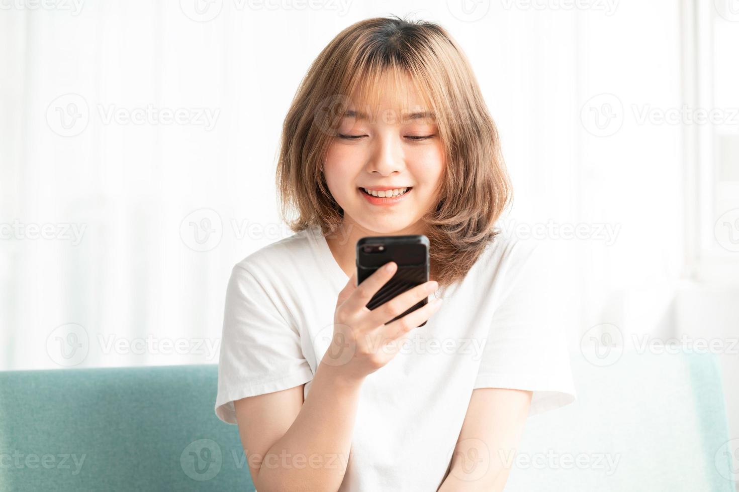 joven asiática sentada en el sofá usando el teléfono con una cara alegre foto