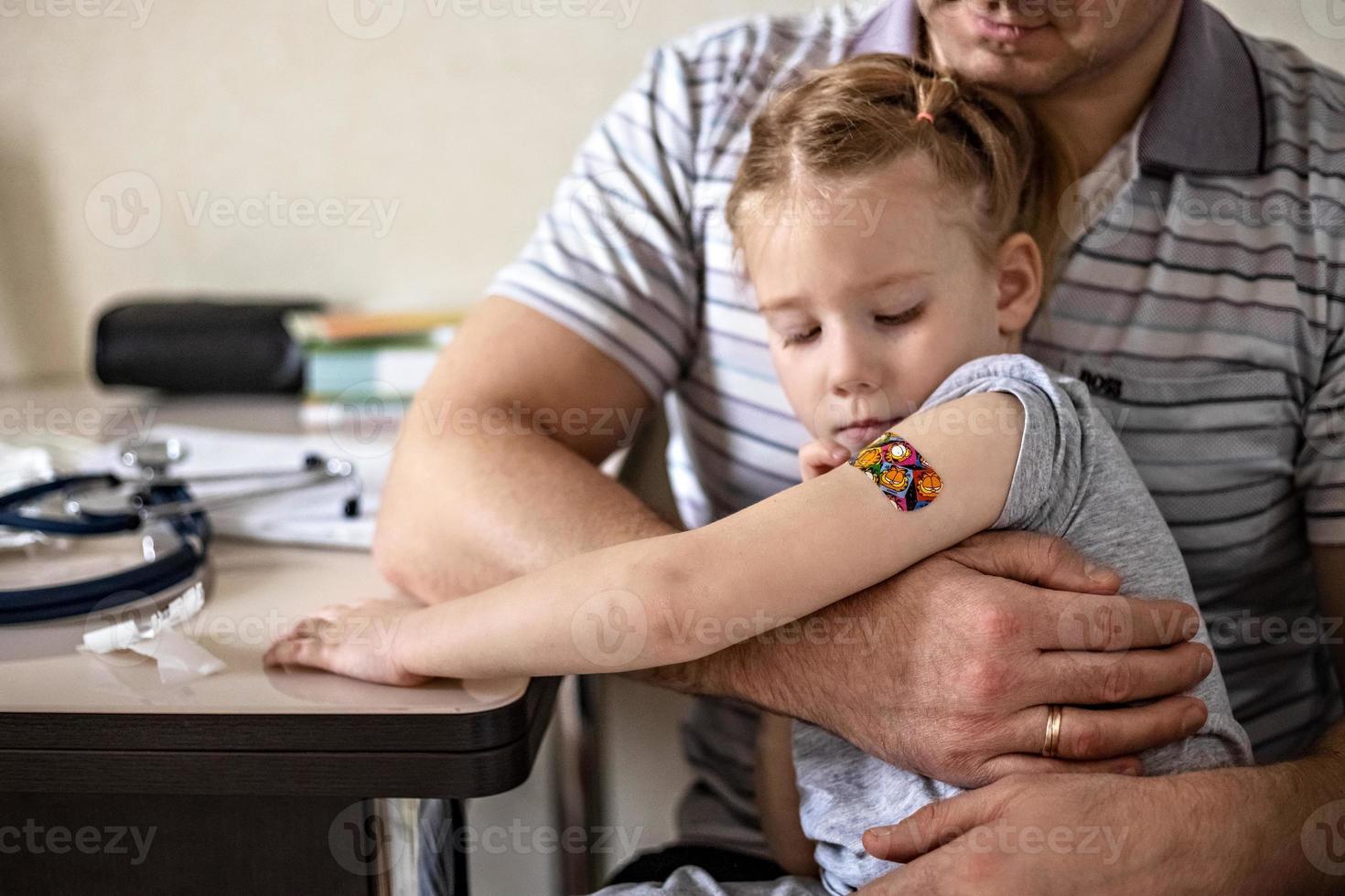 Vaccination of a little girl in her dad's arms in the doctor's office from the coronavirus. Children's funny adhesive plaster. Vaccine against covid-19, flu, infectious diseases. photo