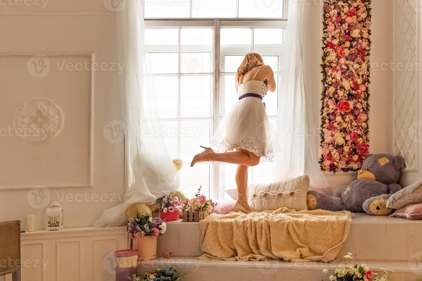 Portrait of a young woman in a white dress against the background of a closed large window, looking out into the street through the glass photo