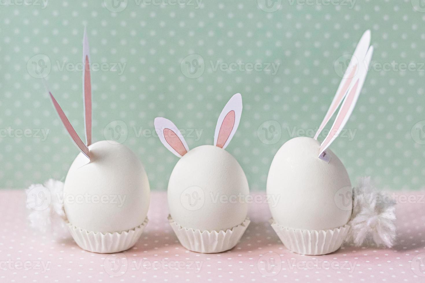 White chicken eggs with bunny ears and tails on bed flowers background. A family. Happy Easter holiday concept photo