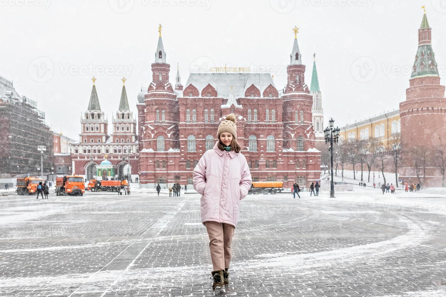 una hermosa joven con una chaqueta rosa camina por la plaza manezhnaya en moscú durante una nevada y una ventisca. los quitanieves están trabajando en segundo plano. foto