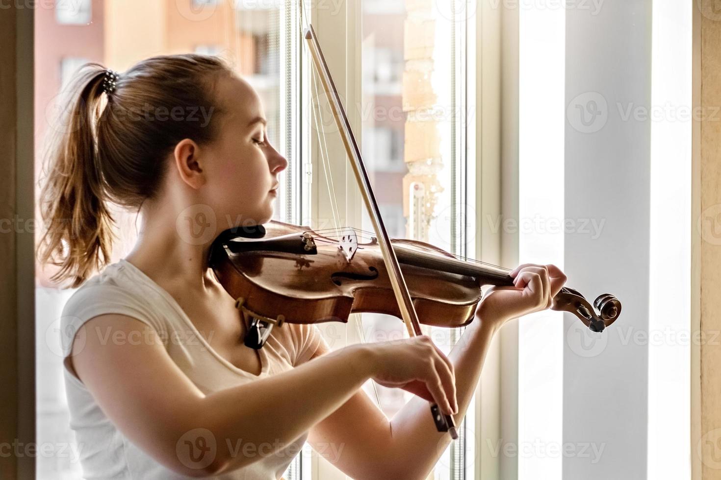 A young girl, a musician, plays the violin on the background of a window photo