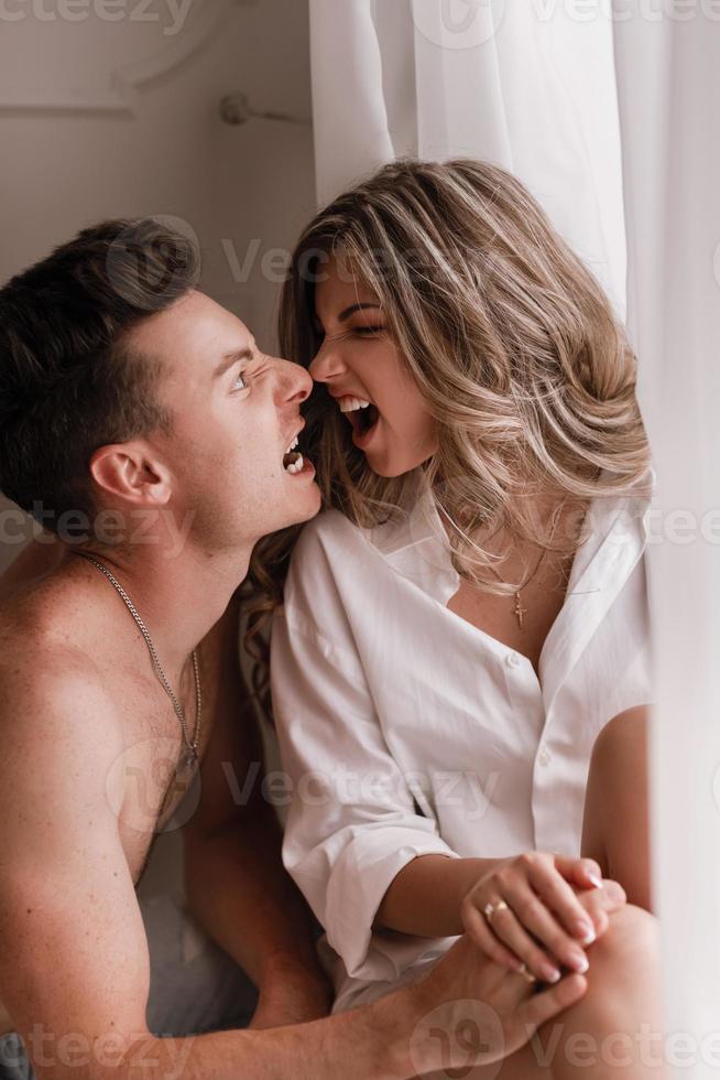 amorosa pareja joven disfrutando de la mañana en casa cerca de la ventana en el día de San Valentín. chica con camisa blanca y chico medio desnudo divirtiéndose juntos foto