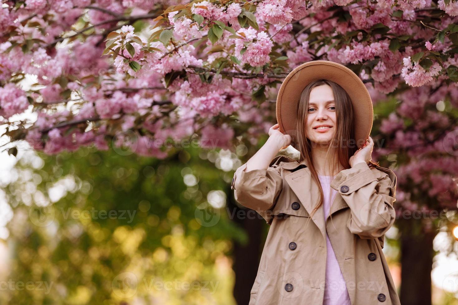 Retrato de joven hermosa chica de moda con sombrero posando cerca de árbol floreciente con flores rosadas en un día soleado foto