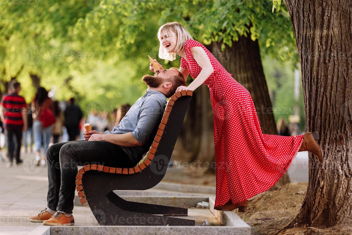 Sonriente pareja de hipsters divirtiéndose y comiendo helado en la ciudad. Elegante joven con barba está sentado en un banco de madera y una mujer rubia en vestido rojo mujer tontea y juega con él foto