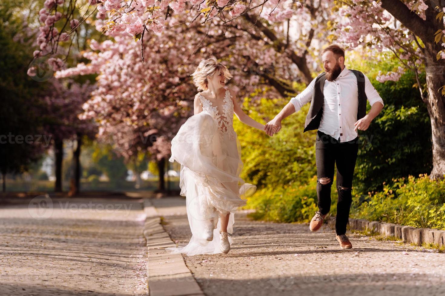 feliz hombre elegante con barba y mujer con vestido largo se divierten en el parque floreciente de la primavera de sakura. pareja de hipsrers recién casados en el parque. recién casados. corre en el parque y toma de la mano. foto