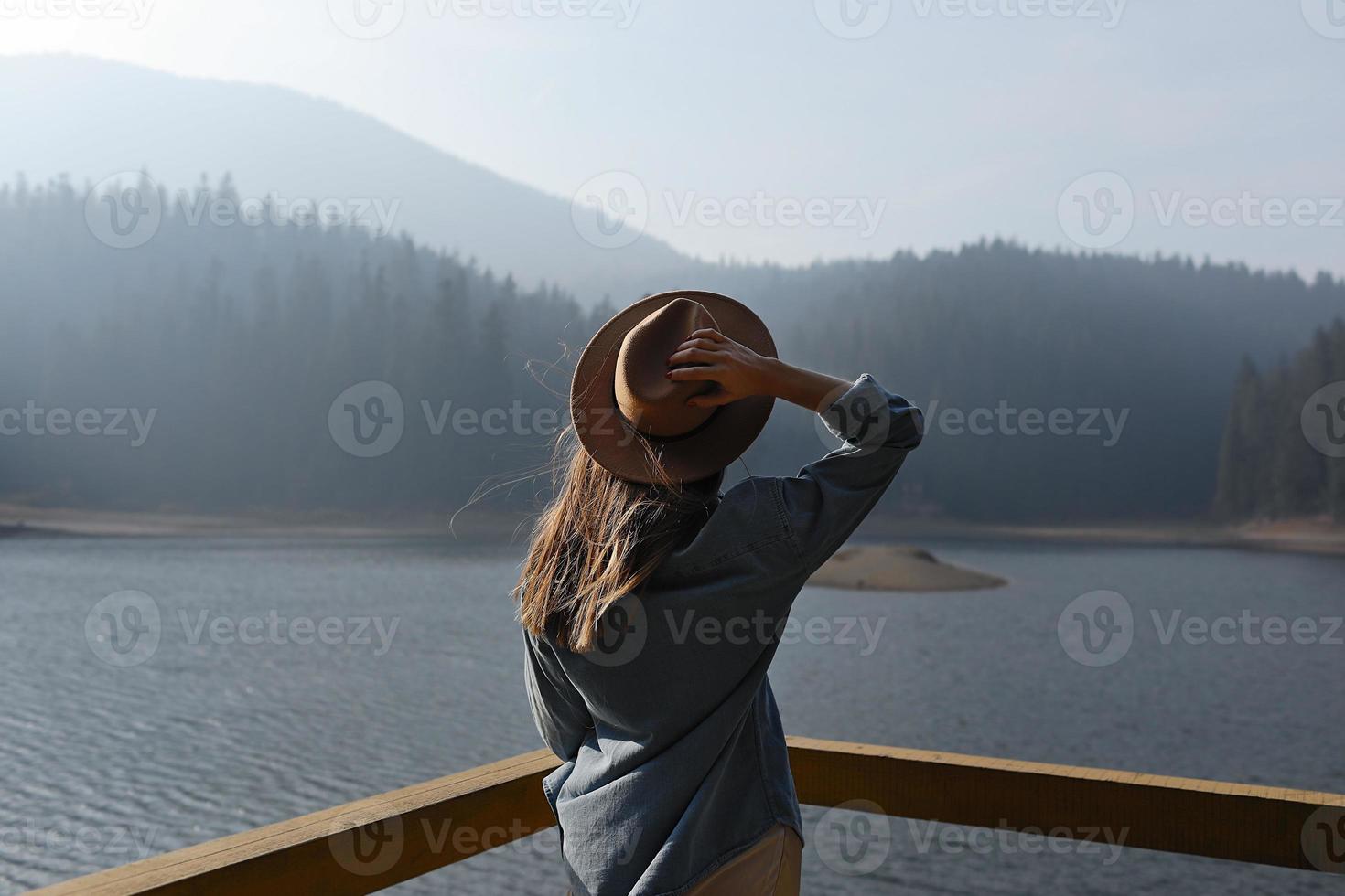 happy young woman in hat enjoys lake view in mountains . Relaxing moments in forest. Back view of stylish girl enjoys the freshness outdoor. Freedom, people, lifestyle, travel and vacation photo