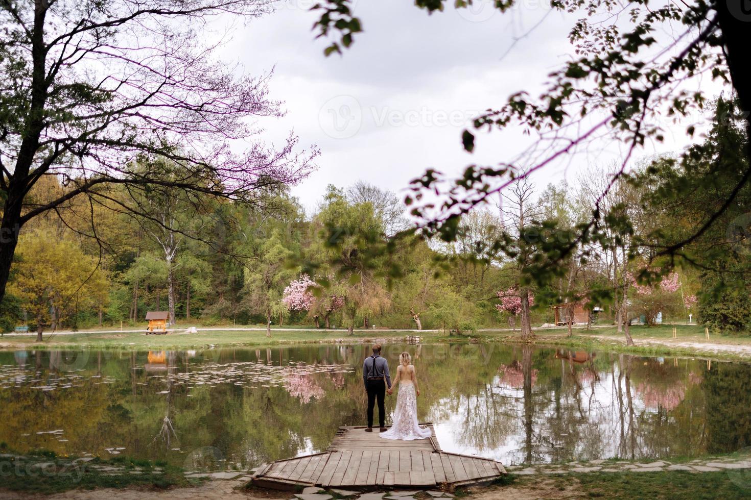 Novio y novia con estilo joven en vestido largo de lujo de pie frente al lago en el parque con flores de cerezo en flor o sakura en el fondo. boda, día de primavera foto