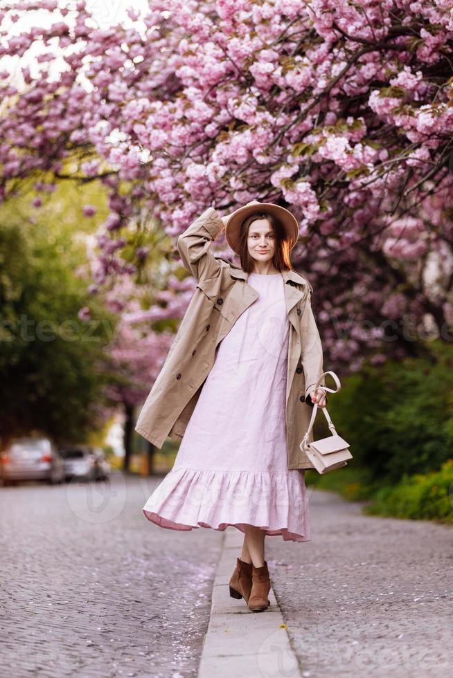 joven hermosa mujer elegante con sombrero y vestido rosa caminando cerca de las flores de sakura en el parque. concepto de primavera. uzhhorod, ucrania foto