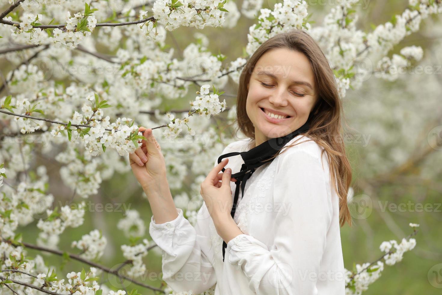 feliz hermosa niña se quitó la máscara médica para respirar el olor de las flores. una niña con una máscara se encuentra en flores. el fin de la cuarentena foto