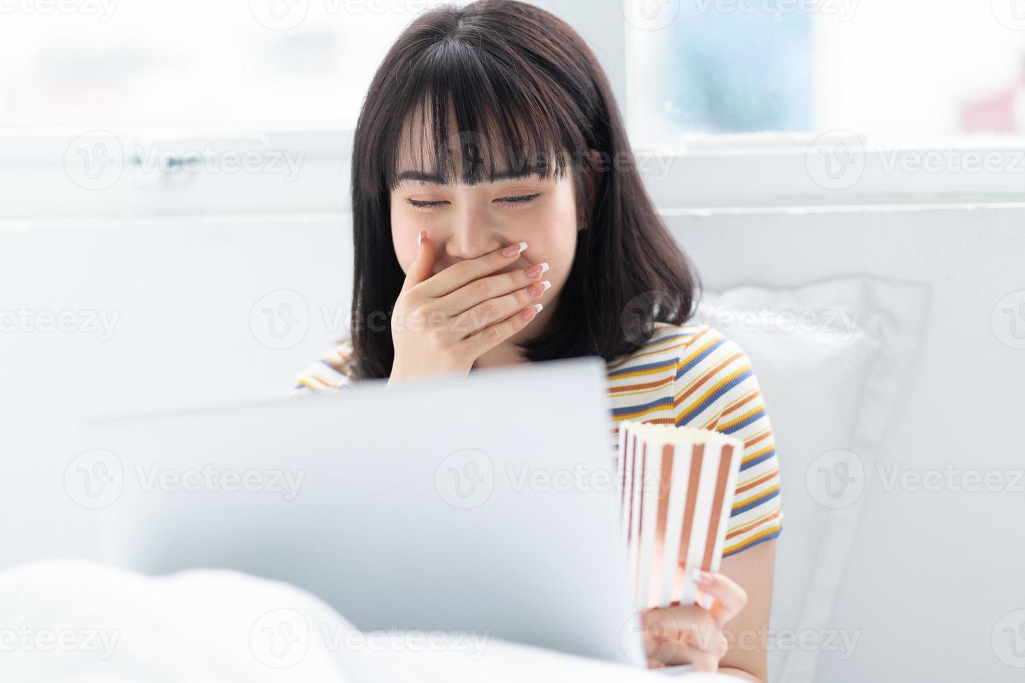 Joven mujer asiática con ordenador portátil para ver programas de televisión, comer palomitas de maíz en casa foto