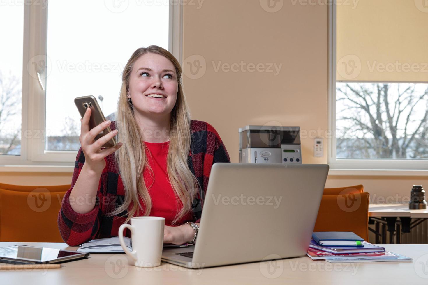 una mujer rubia sonriente sentada en una computadora portátil y hablando por un teléfono celular. foto