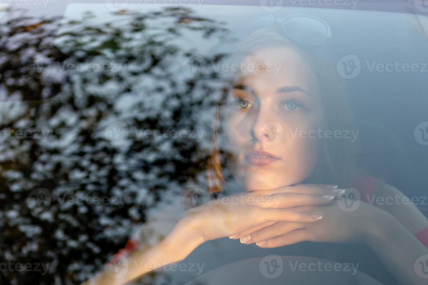 una mujer joven y hermosa con el pelo largo se sienta al volante del automóvil y mira soñadora a través del resplandor del parabrisas delantero. foto