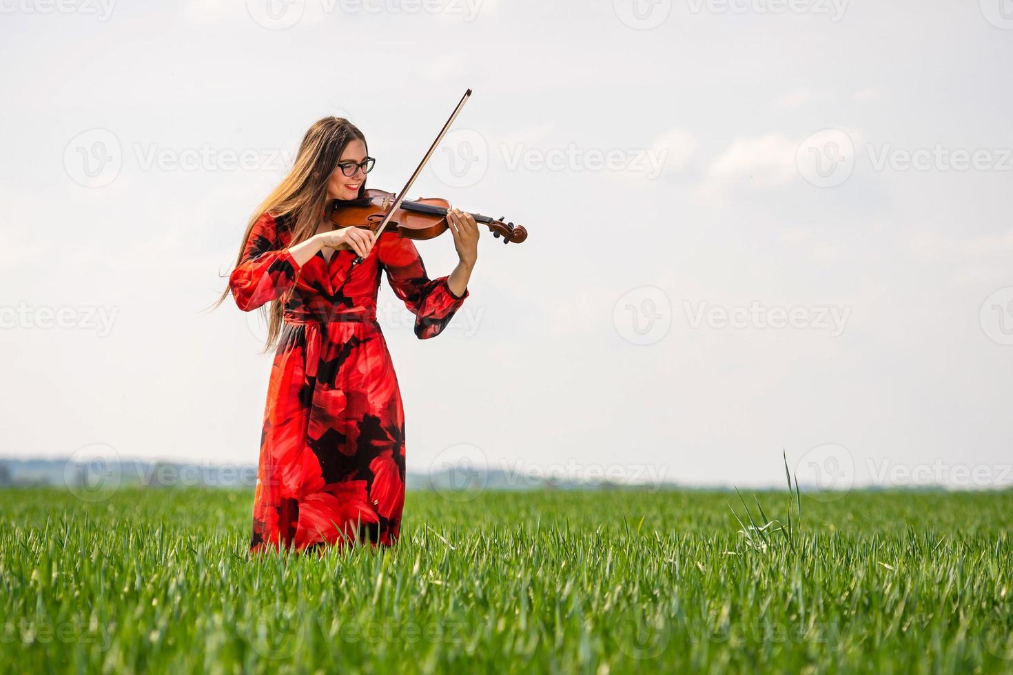 Young woman in red dress playing violin in green meadow - image photo