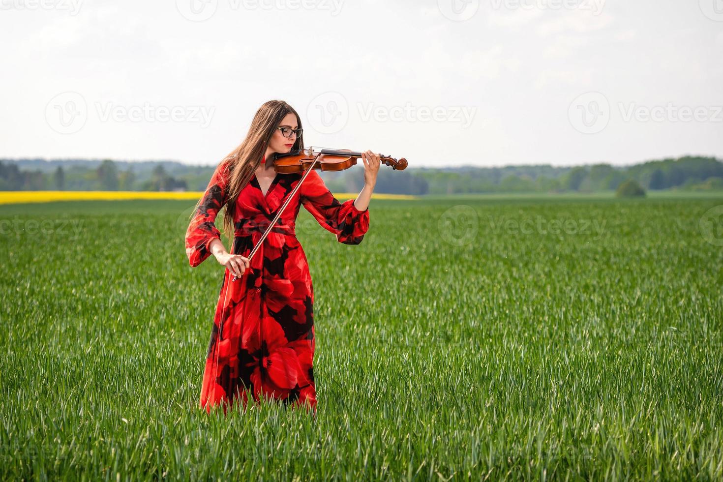 Young woman in red dress playing violin in green meadow - image photo