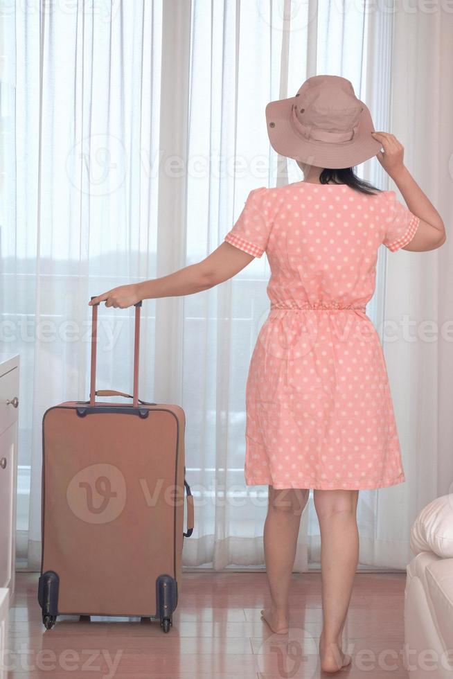 Asian tourist woman in a pink dress standing with her luggage in the hotel bedroom photo