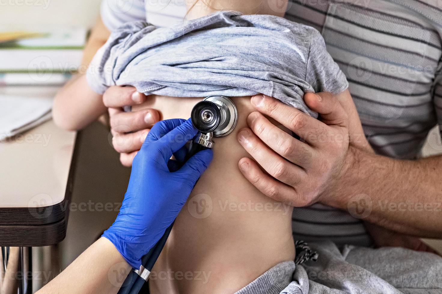 niña en los brazos de su padre en el consultorio del médico en la clínica. el médico examina al niño, escucha los pulmones con un fonendoscopio. tratamiento y prevención de infecciones respiratorias. foto